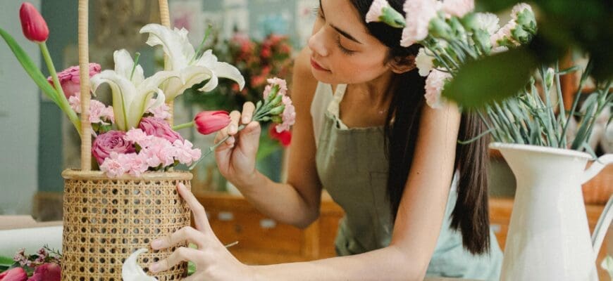 A Woman Arranging Flowers in a Basket on top of a Large Table, with a Vase of Flowers Next to her